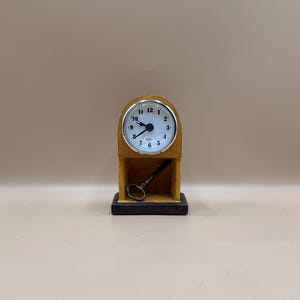 May include: A small, wooden desk clock with a white face and black numerals. The clock has a silver-colored bezel and a key is placed in the clock's base. The clock is set on a black base.