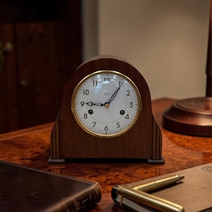 May include: A wooden mantel clock with a white face and gold trim sits on a polished wooden surface. The clock displays the time with black numerals and hands. A leather-bound book and a pen rest nearby.