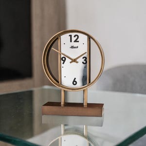 May include: A gold-colored, circular desk clock with a white face and black numerals. The clock has a rectangular wooden base and two vertical supports. The clock face displays the brand name "Kambi".