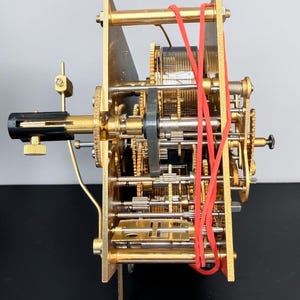 Puede incluir: Un complejo movimiento de reloj mec&aacute;nico con engranajes, ejes y placas de lat&oacute;n. Una banda el&aacute;stica roja asegura algunos componentes. El intrincado dise&ntilde;o muestra el funcionamiento interno de un mecanismo de reloj.