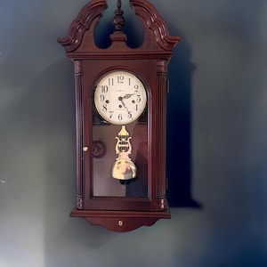 May include: A dark wood wall clock featuring a white circular face with black Arabic numerals and ornate black hands. The clock has a glass-fronted door, fluted side columns, and a decorative pediment top. A gold-toned lyre-shaped pendulum hangs inside.