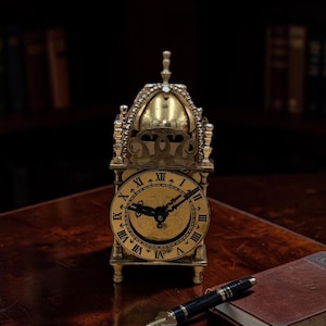 May include: A gold-colored antique clock with Roman numerals on the face, set on a wooden table. A black pen and a small, leather-bound book are next to the clock. The clock has an ornate design and is likely a decorative item.