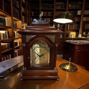 May include: A dark brown wooden mantel clock with a gold-toned face, Roman numerals, and intricate brass corner accents. The clock features a rectangular frame with a decorative top finial and four smaller finials at the corners of the base.