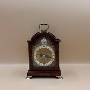 May include: A dark wood antique clock with a handle. The clock face features Roman numerals and decorative elements. The clock is supported by silver-coloured feet.