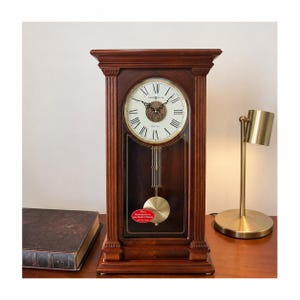 May include: A wooden Howard Miller clock with a cream-colored face and Roman numerals. The clock has a pendulum and is set in a dark brown wooden frame. A gold-colored lamp is to the right of the clock.