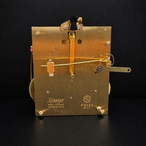 Puede incluir: Un movimiento de reloj de lat&oacute;n con intrincados engranajes y palancas. La marca Kieninger es visible, junto con el texto "Made in Germany". El movimiento es rectangular y tiene un dise&ntilde;o mec&aacute;nico complejo.