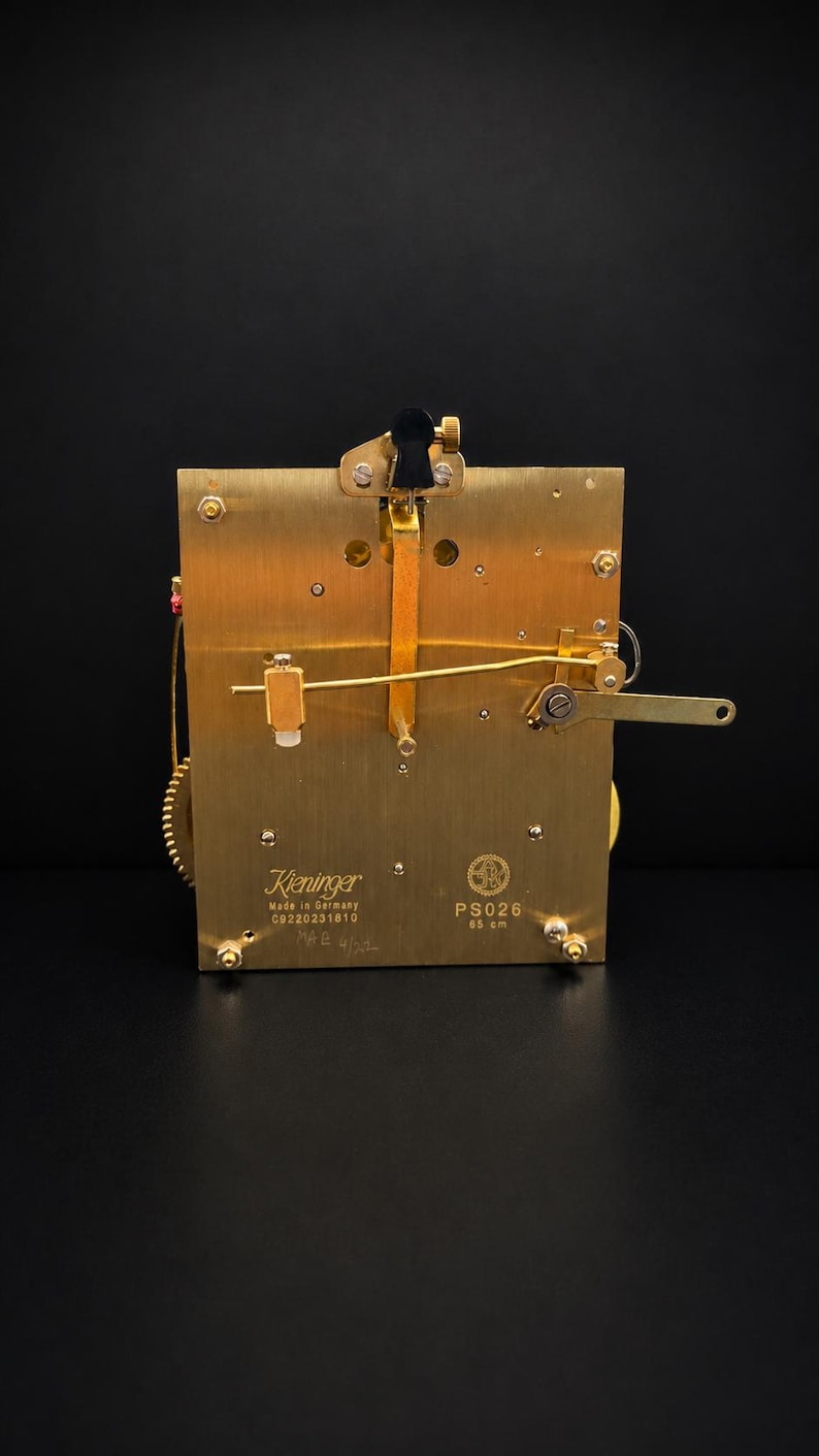 Puede incluir: Un movimiento de reloj de lat&oacute;n con intrincados engranajes y palancas. La marca Kieninger es visible, junto con el texto "Made in Germany". El movimiento es rectangular y tiene un dise&ntilde;o mec&aacute;nico complejo.