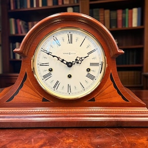 May include: A wooden mantel clock with a cream-colored face and black Roman numerals. The clock has a curved top and a rectangular base. The clock is made of reddish-brown wood and sits on a wooden table. The clock face has black hands and the brand name "Howard Miller".
