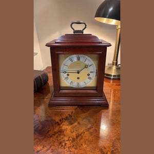 May include: A rectangular, wooden mantel clock with a brass handle. The clock face is cream-colored with black Roman numerals and hands. The clock is set on a wooden surface.