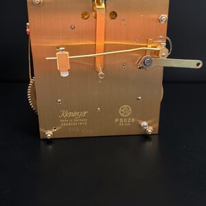 Puede incluir: Un movimiento de reloj de lat&oacute;n con engranajes y palancas intrincados. La marca Kieninger es visible, junto con el texto "Made in Germany". El movimiento tiene forma rectangular con varios componentes.