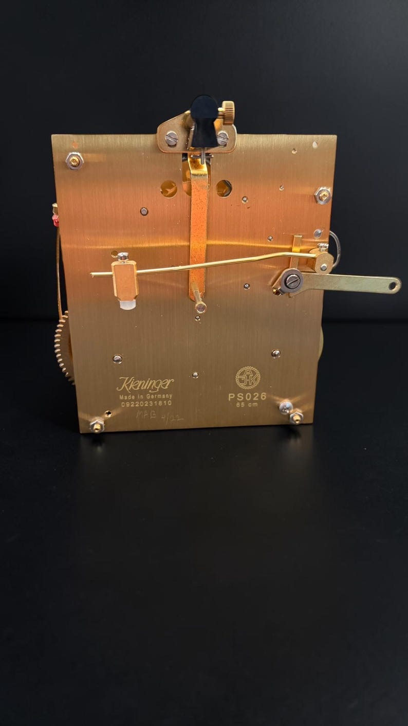 Puede incluir: Un movimiento de reloj de lat&oacute;n con engranajes y palancas intrincados. La marca Kieninger es visible, junto con el texto "Made in Germany". El movimiento tiene forma rectangular con varios componentes.