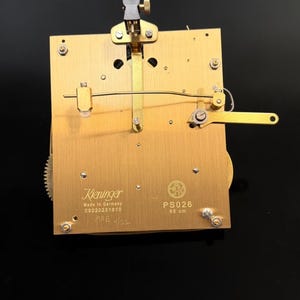 Puede incluir: Un movimiento de reloj de lat&oacute;n con engranajes y palancas intrincados. El movimiento est&aacute; marcado "Kieninger Made in Germany" y "PS026 85 cm". La imagen muestra una vista detallada de los mecanismos internos del reloj.