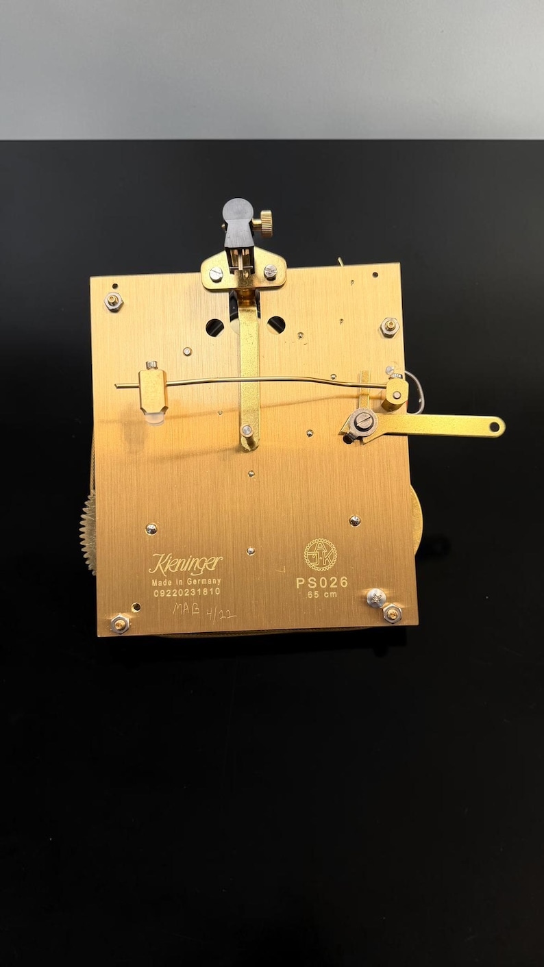 Puede incluir: Un movimiento de reloj de lat&oacute;n con engranajes y palancas intrincados. El movimiento est&aacute; marcado "Kieninger Made in Germany" y "PS026 85 cm". La imagen muestra una vista detallada de los mecanismos internos del reloj.