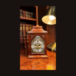 May include: A wooden mantel clock with a detailed face and Roman numerals. The clock has a decorative design and is set on a wooden surface. The clock is illuminated by a brass lamp.