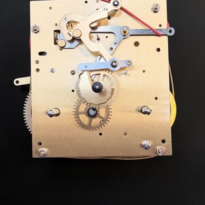 Puede incluir: Primer plano de un mecanismo de reloj con componentes de lat&oacute;n y plata. Se ven engranajes, palancas y muelles, junto con un cable rojo. El mecanismo est&aacute; montado sobre una placa rectangular de lat&oacute;n.