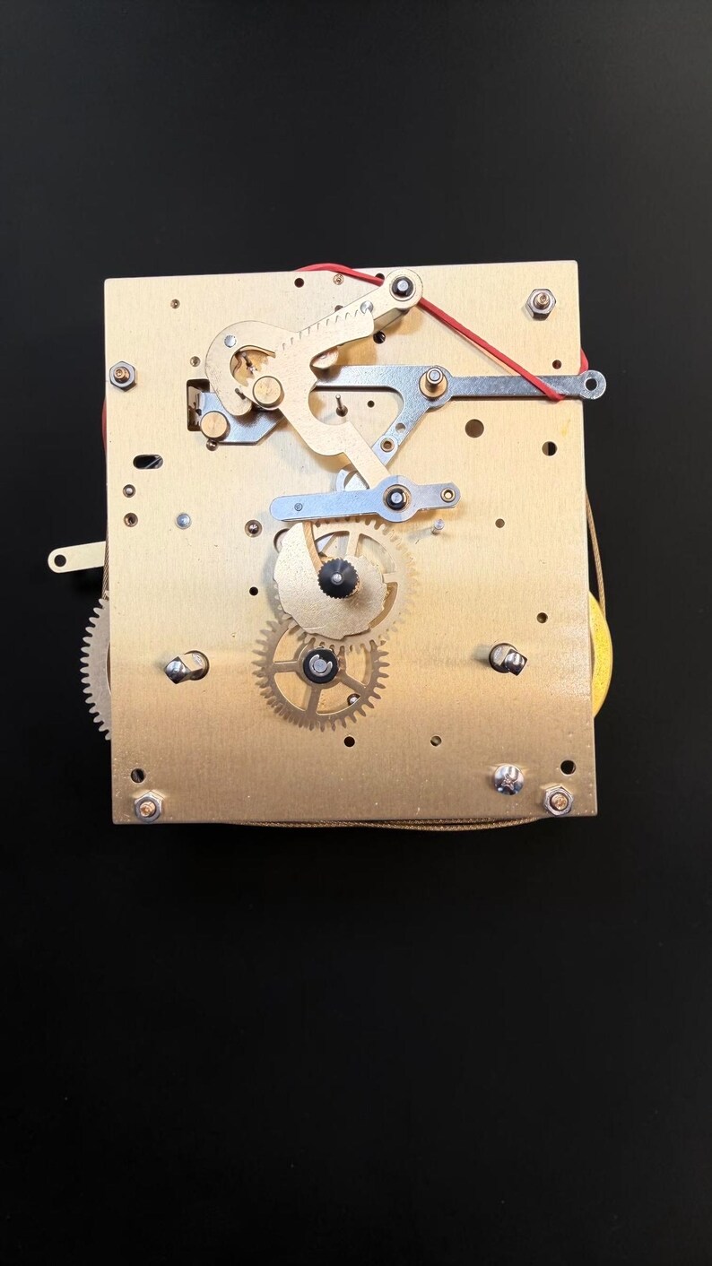 Puede incluir: Primer plano de un mecanismo de reloj con componentes de lat&oacute;n y plata. Se ven engranajes, palancas y muelles, junto con un cable rojo. El mecanismo est&aacute; montado sobre una placa rectangular de lat&oacute;n.