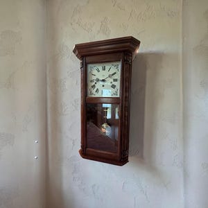 May include: A dark wood wall clock with a rectangular shape. The clock face has Roman numerals and decorative hands. A glass panel reveals the pendulum. The clock is mounted on a textured wall.