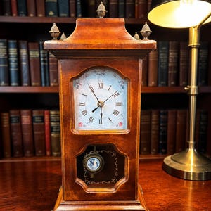 May include: A wooden antique clock with a light-colored face featuring Roman numerals and delicate floral accents. The clock has a pendulum visible through a small window and is adorned with decorative finials. The clock is set on a wooden surface.