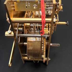 Puede incluir: Primer plano de un mecanismo de reloj mec&aacute;nico con engranajes, placas y un resorte helicoidal de lat&oacute;n. Bandas de goma rojas aseguran algunos componentes. El dise&ntilde;o intrincado muestra el interior de un reloj.
