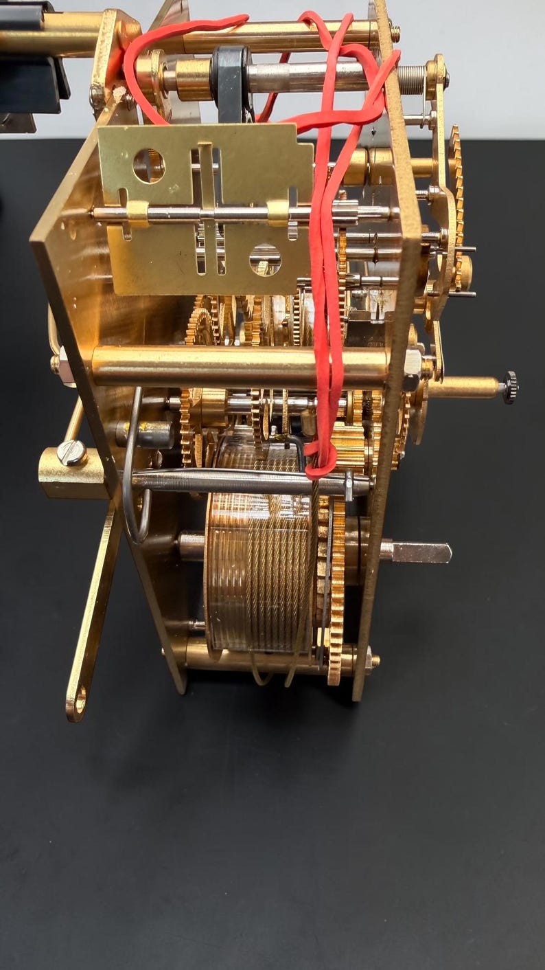 Puede incluir: Primer plano de un mecanismo de reloj mec&aacute;nico con engranajes, placas y un resorte helicoidal de lat&oacute;n. Bandas de goma rojas aseguran algunos componentes. El dise&ntilde;o intrincado muestra el interior de un reloj.