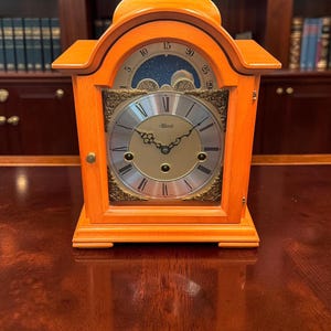 May include: An orange wooden mantel clock with a gold-toned face, black Roman numerals, and ornate black hands. The clock features a moon phase dial at the top with numbers 1 to 29, a gold handle on top, and the brand name Hermle printed on the face.