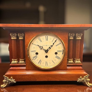May include: A rectangular wooden mantel clock with a warm brown finish. The clock face is cream-colored with black Roman numerals and ornate black hands. It features gold-toned decorative columns on each side and intricate gold-toned feet.