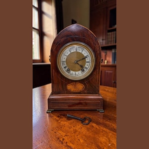 May include: An antique wooden mantel clock with a round face and Roman numerals. The clock has a dark brown finish with decorative inlay and a key is resting on the wooden surface in front of it. The clock is a vintage item.