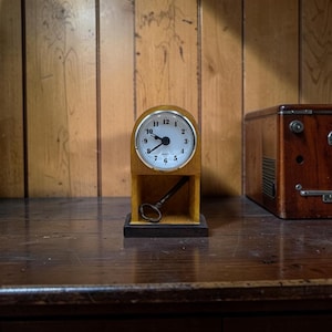 May include: A small, wooden desk clock with a white face and black numerals. The clock has a gold-colored metal bezel and a key resting in the base. A wooden box is to the right. The clock is on a dark wooden surface.