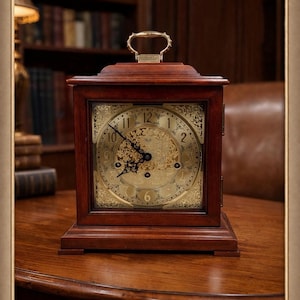 Peut inclure: Une horloge de cheminée rectangulaire en bois avec un cadran doré et une poignée sur le dessus. Le cadran de l'horloge comporte des chiffres romains et des détails décoratifs. L'horloge est posée sur une table en bois.