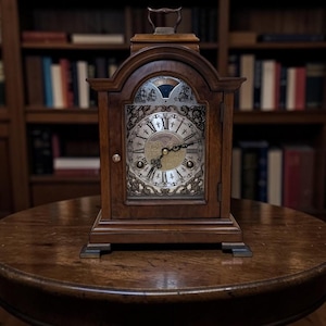 May include: A wooden antique clock with a decorative face and Roman numerals, set on a round wooden table. The clock has a handle on top and is surrounded by a dark wooden bookshelf in the background.