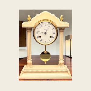 May include: A cream-colored mantel clock with a gold-toned clock face and pendulum. The clock has Roman numerals and black hands. The clock is supported by two cream-colored columns and topped with gold finials. The clock sits on a wooden surface.