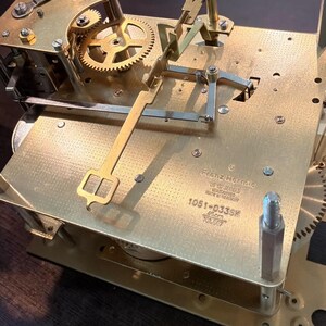 Puede incluir: Primer plano de un mecanismo de reloj de lat&oacute;n con engranajes, palancas y un sistema de p&eacute;ndulo. El mecanismo del reloj tiene el texto "Franz Hermle" y "1051-033SK" impreso. El mecanismo del reloj mide 45 cm de largo.