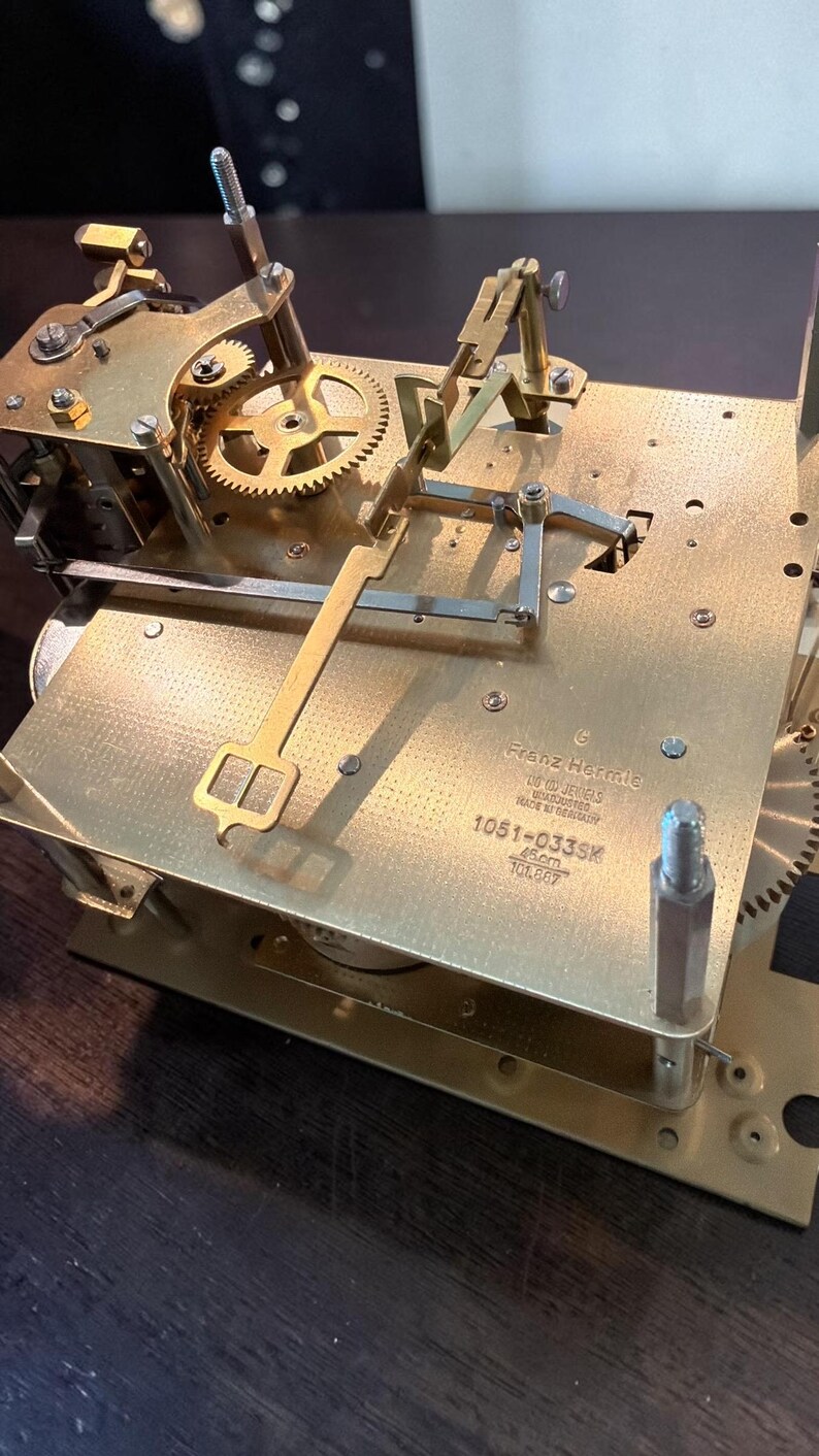 Puede incluir: Primer plano de un mecanismo de reloj de lat&oacute;n con engranajes, palancas y un sistema de p&eacute;ndulo. El mecanismo del reloj tiene el texto "Franz Hermle" y "1051-033SK" impreso. El mecanismo del reloj mide 45 cm de largo.