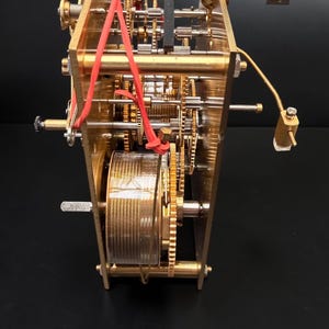 Puede incluir: Vista detallada de un mecanismo de reloj con engranajes de lat&oacute;n, muelles y un componente cil&iacute;ndrico. Bandas rojas aseguran partes del mecanismo. El intrincado dise&ntilde;o se presenta sobre un fondo oscuro, mostrando la precisi&oacute;n del reloj.