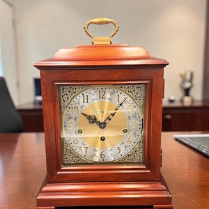 May include: A rectangular wooden clock with a gold-colored handle. The clock face is gold with black Roman numerals and ornate detailing. The clock is made of dark brown wood and sits on a matching base.