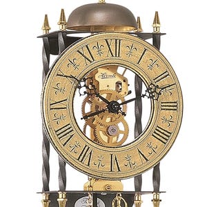 May include: A gold and black Hermle clock with Roman numerals on the clock face. The clock has a decorative design with gold accents and a pendulum. The clock's gears are visible through the center of the clock face.