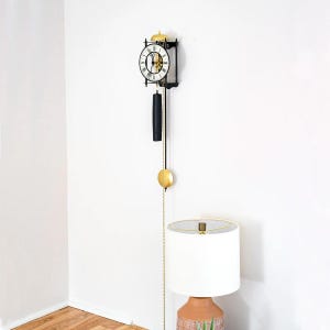 May include: A black and gold wall clock with a round face and Roman numerals. The clock has a pendulum and a gold chain. The clock is hanging on a white wall. The clock is approximately 24 inches long.