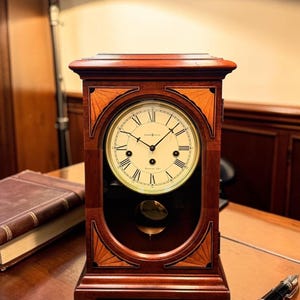 May include: A wooden mantel clock with a cream-coloured face, Roman numerals, and a pendulum. The clock is housed in a rectangular, dark wood case with decorative inlays. A pen and a book are on the table.