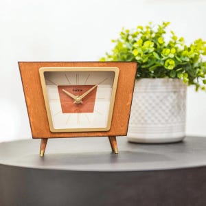 May include: A wooden clock with a mid-century modern design. The clock has a square face with a gold-colored frame and a light brown wooden body. The clock face has a white background with a red-brown square in the center. The clock is labeled "Hermie".