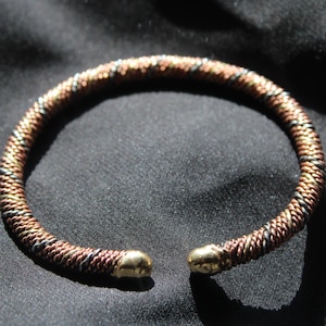 May include: A woven cuff bracelet in shades of copper, bronze, and silver. The bracelet has a rounded shape with gold-coloured caps on each end. The jewellery is displayed on a dark, textured surface.