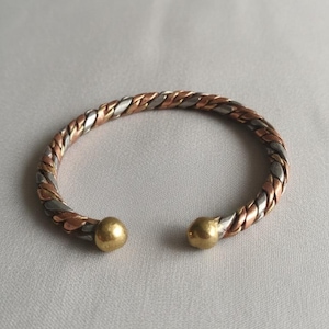 Puede incluir: Una pulsera de brazalete trenzada con tonos de cobre, plata y oro. El diseño abierto presenta una bola dorada en cada extremo. La pulsera se muestra sobre una superficie gris claro.