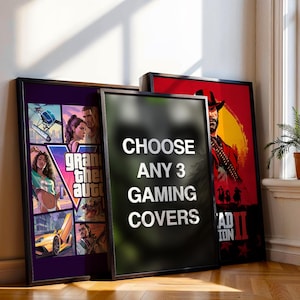 May include: Three framed rectangular posters leaning against a wall. The left poster features Grand Theft Auto V artwork. The centre poster is black with white text reading CHOOSE ANY 3 GAMING COVERS. The right poster displays Red Dead Redemption 2.