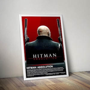 May include: A framed poster for the video game "Hitman: Absolution." The poster features a bald man in a black coat, viewed from the back, holding two pistols. The title is prominently displayed in white letters against a red background.
