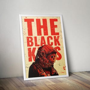 May include: Framed art print featuring a red and black illustration of a space helmet with the text "THE BLACK KISS" in bold red letters. The artwork has a vintage, distressed look, with a cream-colored background and a white frame.