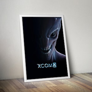 Puede incluir: Un póster enmarcado con un primer plano de una cara alienígena amenazante con dientes afilados y ojos brillantes, emergiendo de la oscuridad. El póster tiene el texto "XCOM 2" en una fuente futurista, sobre un fondo negro.