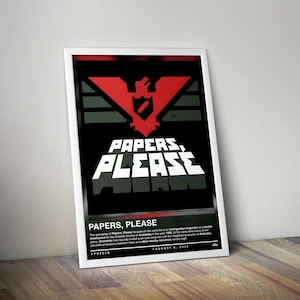 May include: A framed poster for the video game "Papers, Please." The poster features a red emblem over the words "PAPERS, PLEASE" in white block letters against a black background. The poster is leaning against a wooden surface.