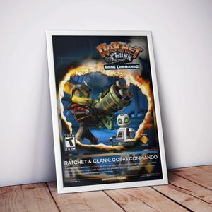 May include: A framed poster for the video game "Ratchet & Clank: Going Commando." The artwork features the game's characters, Ratchet and Clank, with a large green weapon. The poster has a white frame and is leaning against a wall.
