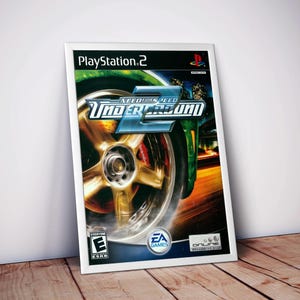 May include: Framed poster for the video game Need for Speed: Underground 2. The image shows a close-up of a car wheel with a gold rim and red brake calipers. The game's logo is displayed, along with the PlayStation 2 logo and the ESRB rating.