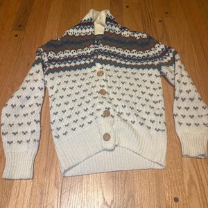 May include: Cream-colored cardigan sweater with a patterned yoke in shades of brown, teal, and burgundy. The sweater features a button-up front and a repeating heart-shaped pattern. The sleeves are long and the collar is a shawl style.