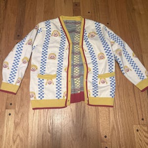 May include: Cream-colored cardigan sweater with a repeating pattern of smiling faces and blue checkered stripes. The sweater has yellow trim, red accents, and two front pockets. The interior lining features a checkered pattern.